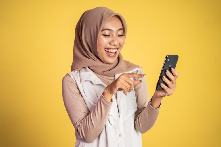 Young asian muslim woman laughing at her smartphone isolated on yellow background