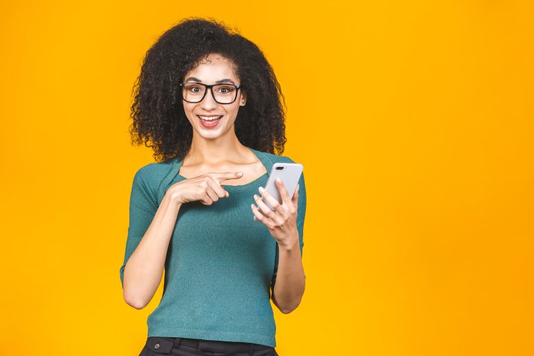 Happy amazed shocked young african american woman casually dressed standing isolated over yellow background, holding mobile phone.